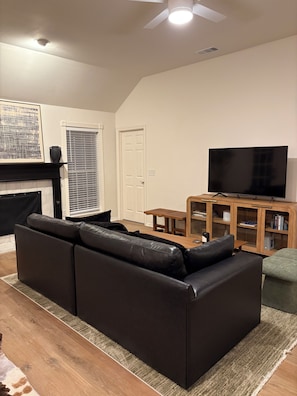 A living room with a black leather sofa, a wooden coffee table, a flat-screen TV, a fireplace, and a bookshelf. - Spacious 3BR House in Frisco - Minutes from Top Event Attractions (Frisco)