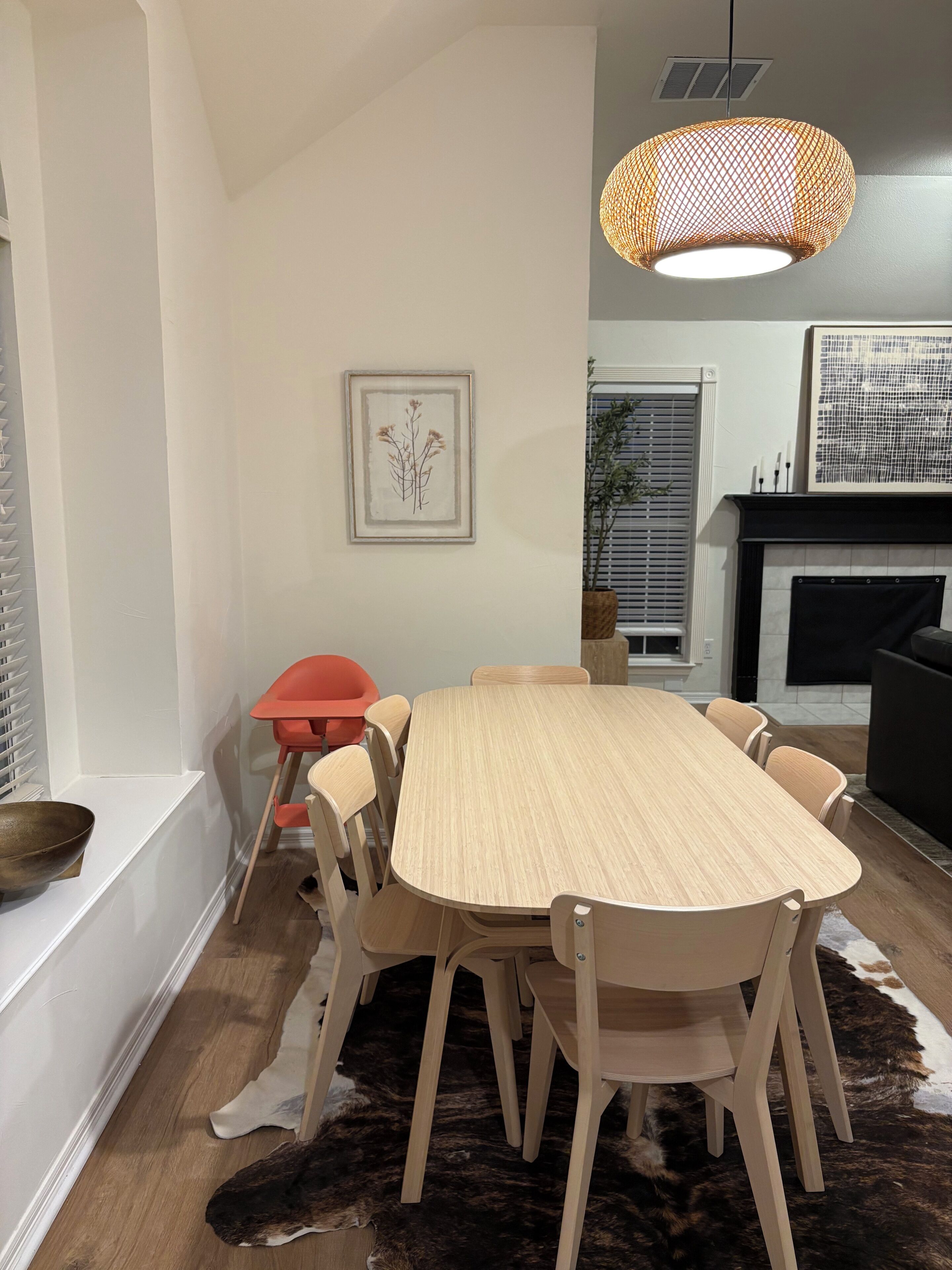 A dining area with a round wooden table, six chairs, a hanging light fixture, a framed picture on the wall, and a fur rug on the floor.