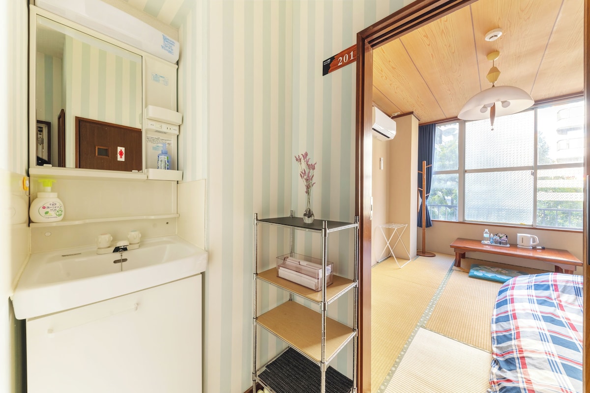 Quarto de hotel com parede listrada, porta de madeira, prateleira com artigos de higiene pessoal, mesa de cabeceira com abajur e janela com vista.