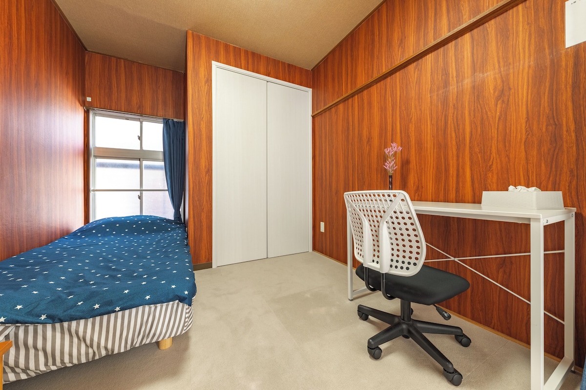 Quarto com cama, escrivaninha, cadeira e janela com cortinas.