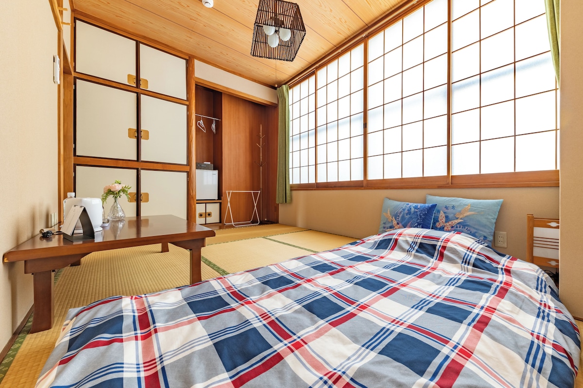 Um quarto japonês tradicional com tatames, uma cama com colcha xadrez, uma escrivaninha de madeira e portas shoji.
