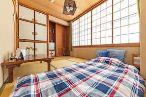 Um quarto japonês tradicional com tatames, uma cama com colcha xadrez, uma escrivaninha de madeira e portas shoji.