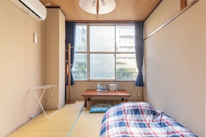 Quarto pequeno com cama, mesa e chaleira, e janela com vista para prédios.