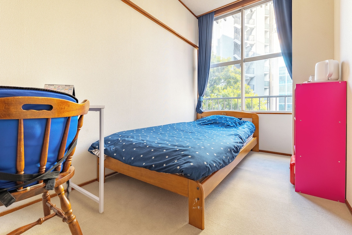 Quarto pequeno com uma cama de solteiro, estrutura de madeira, colcha azul, cadeira de madeira, escrivaninha branca, armário rosa e janela com cortinas.