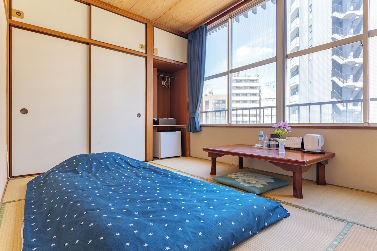 Quarto japonês tradicional com tatames, futon, mesa de madeira com um vaso e vista para a cidade através de janelas amplas.