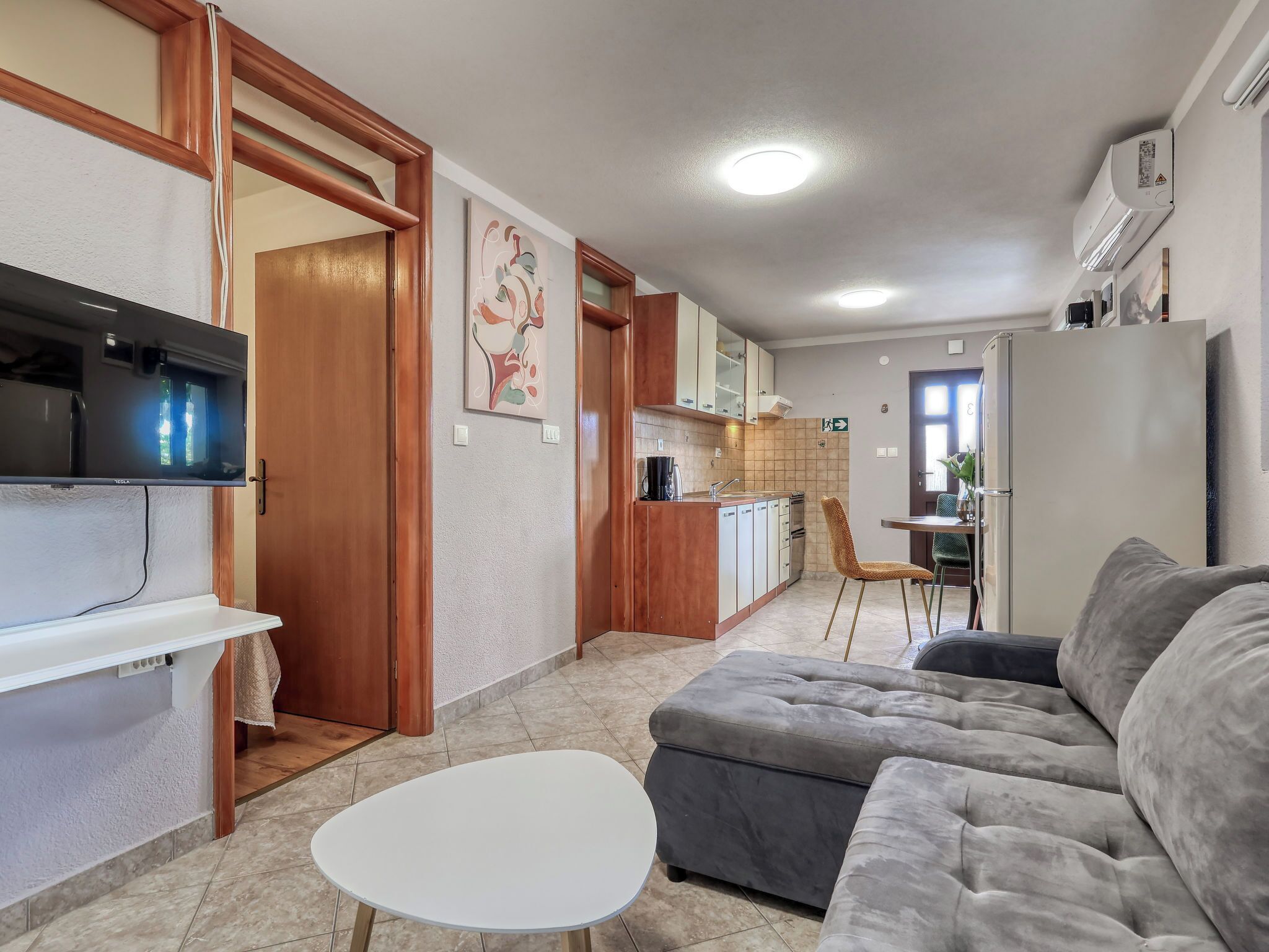 Een functionele leefruimte met een bank, een eettafel, een kitchenette met kastjes en een spoelbak, en een televisie aan de wand.