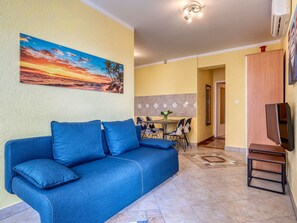 Ein Wohnzimmer mit blauem Sofa, einem Essbereich mit Tisch und Stühlen sowie einer Küche im Hintergrund.