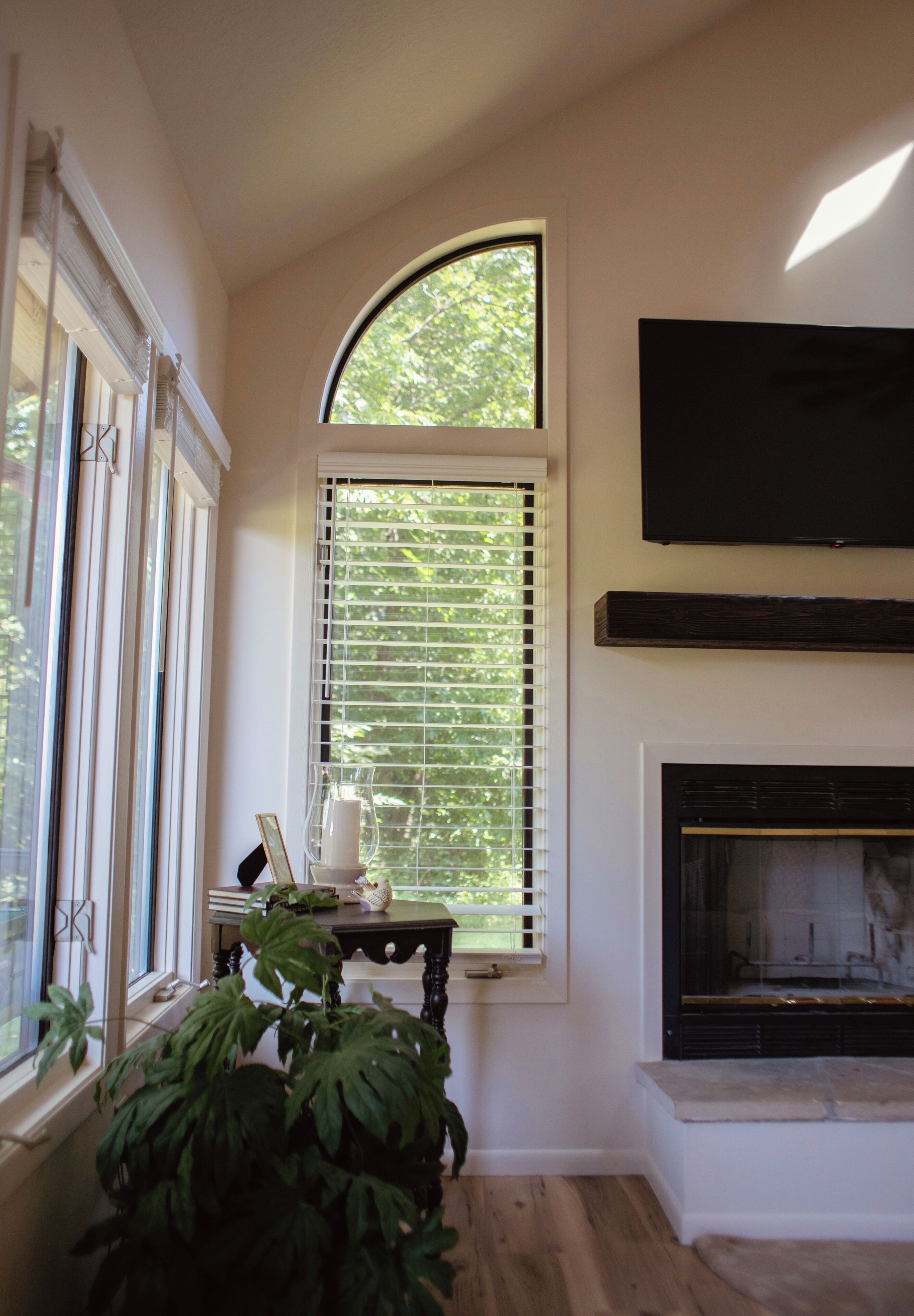 A room with a fireplace, a large window with blinds, and a television mounted above it.