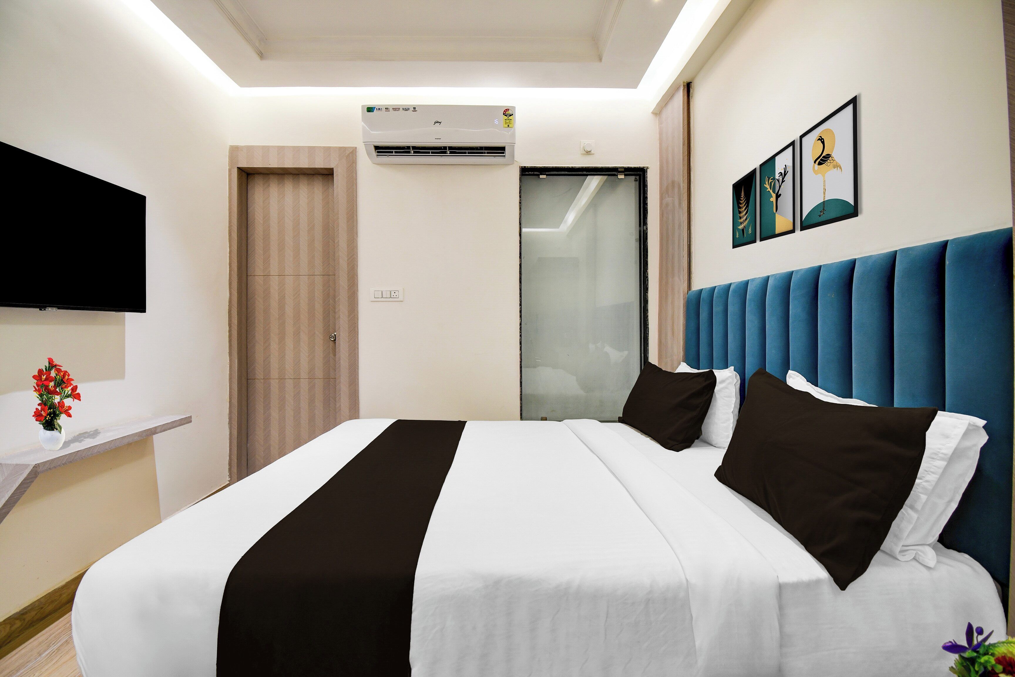 A hotel room with a bed, a flat-screen TV, an air conditioning unit, a wall-mounted artwork, and a window with blinds.