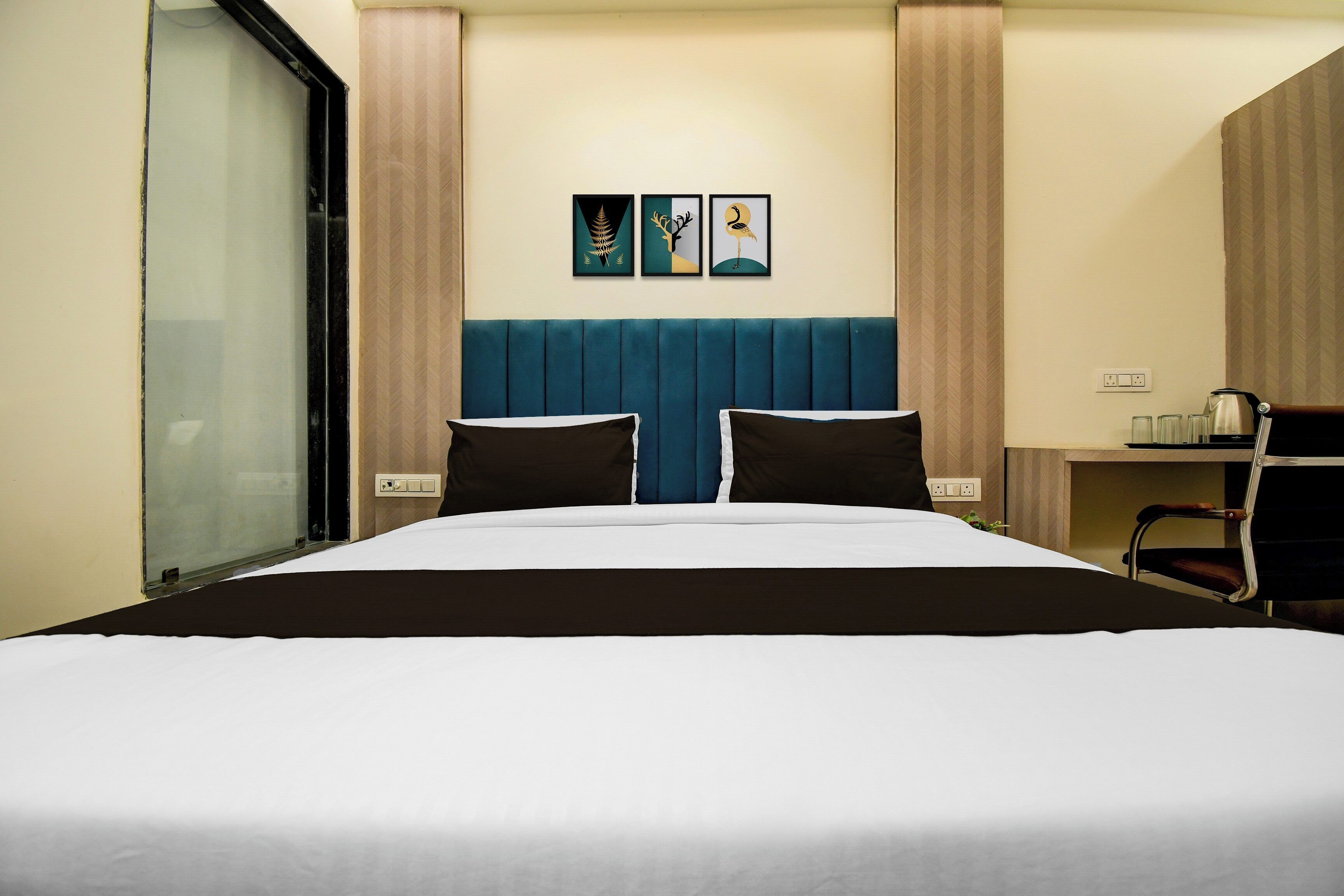 Hotellihuone, jossa on suuri sänky, pääty, kaksi seinälle kiinnitettyä taideteosta, työpöytä tuolilla ja ikkuna, jossa on sälekaihtimet.