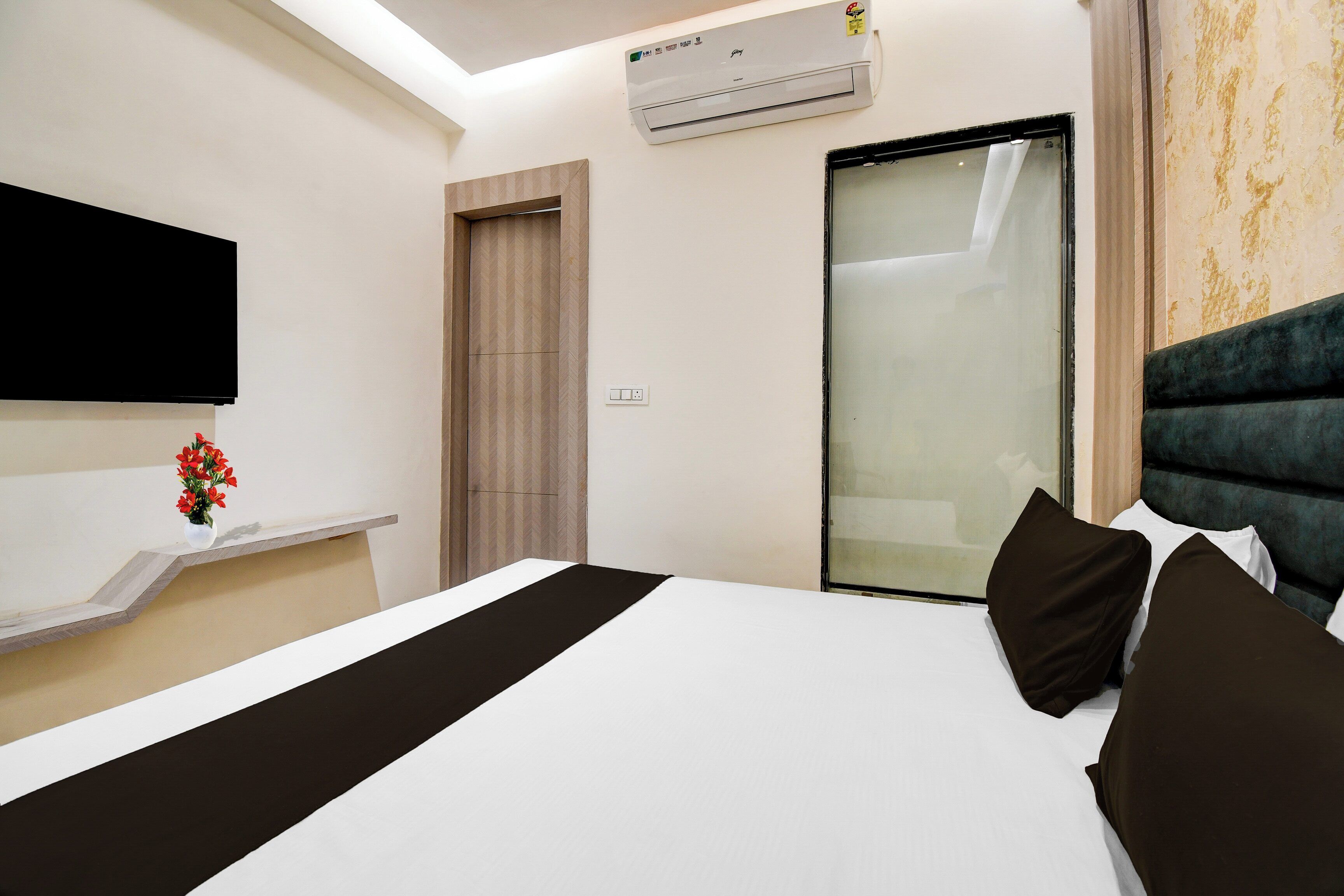 A hotel room with a bed, a flat-screen TV, an air conditioning unit, a sliding glass door, and a small vase with flowers on a shelf.