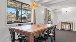 A dining area with a wooden table, black chairs, a woven pendant light, and a view of trees through large windows. - Beach Street Bungalow (Vincentia)