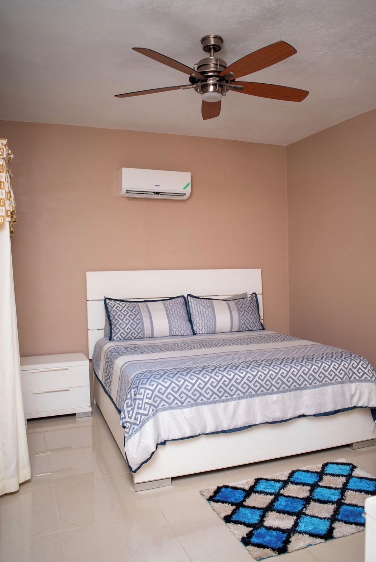 A bedroom with a bed, nightstand, air conditioning unit, ceiling fan, and a patterned rug.