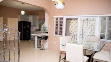 A dining area with a glass table and chairs, a kitchen in the background, and a staircase leading to another level.