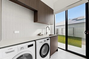 A modern kitchen with a washing machine, a sink, and a large sliding glass door leading to a backyard. - Luxe Harbour Breeze at Newport (Newport)