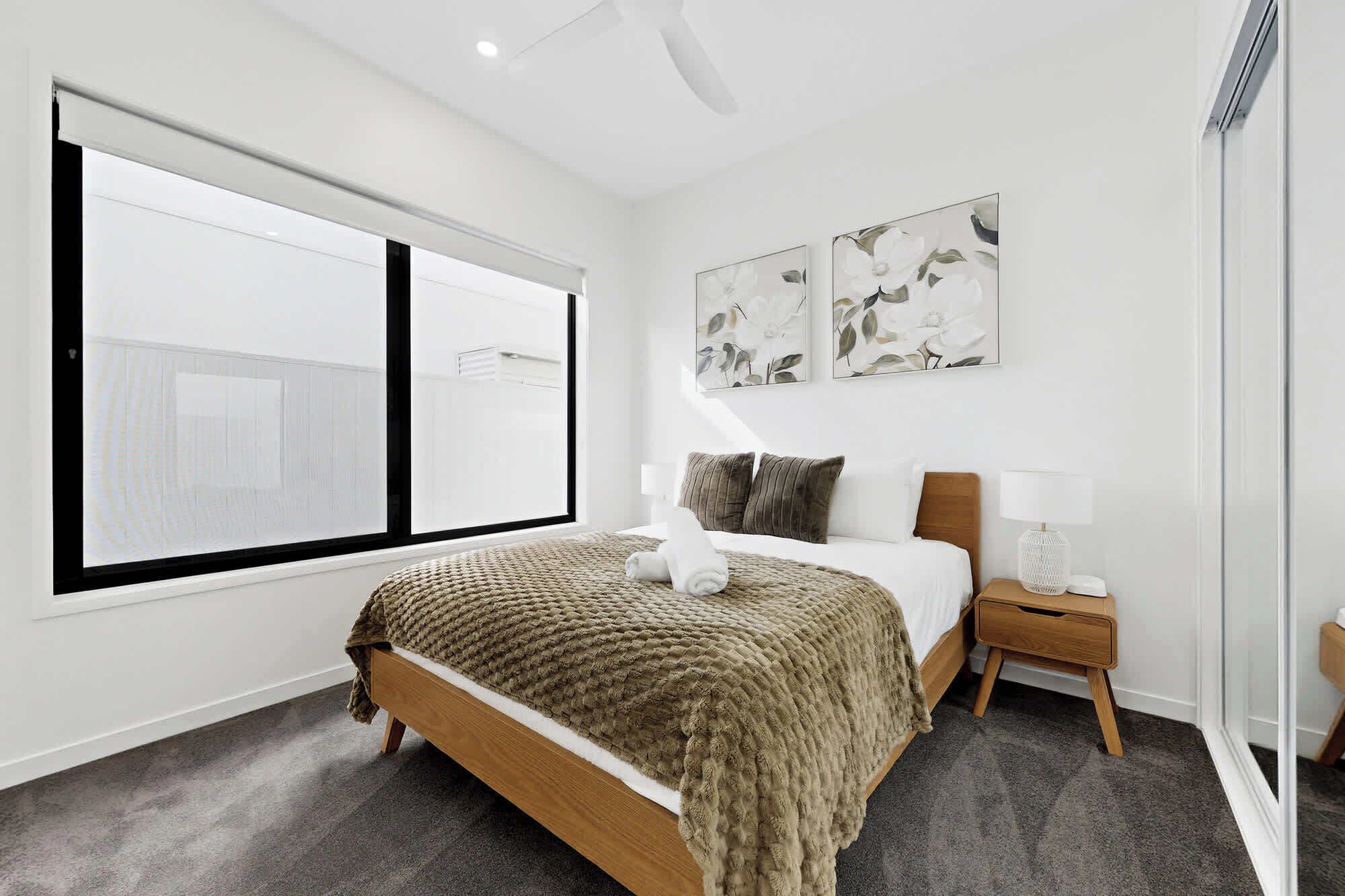 Una habitación de hotel moderna con un ventanal grande, una cama con una manta marrón, dos obras de arte en la pared, una mesita de noche con una lámpara y una pequeña mesita auxiliar de madera.