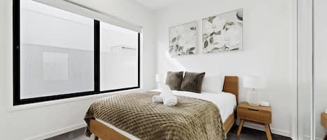 Una habitación de hotel moderna con un ventanal grande, una cama con una manta marrón, dos obras de arte en la pared, una mesita de noche con una lámpara y una pequeña mesita auxiliar de madera.