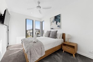 A modern bedroom with a large bed, a ceiling fan, a nightstand, and a window with a view of buildings. - Luxe Harbour Breeze at Newport (Newport)