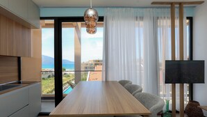 A modern living space with a wooden dining table, grey upholstered chairs, a flat-screen TV, and a view of a coastal area. - Majestic View Apartment Palm (Orikum)