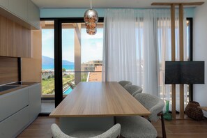 A modern living space with a wooden dining table, grey upholstered chairs, a flat-screen TV, and a view of a coastal area. - Majestic View Apartment Palm (Orikum)