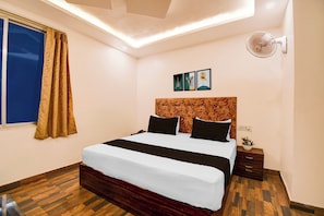 A hotel room with a bed, a nightstand, a wall-mounted fan, and a window with curtains. - Townhouse Civil Lines Prayagraj (Prayagraj)