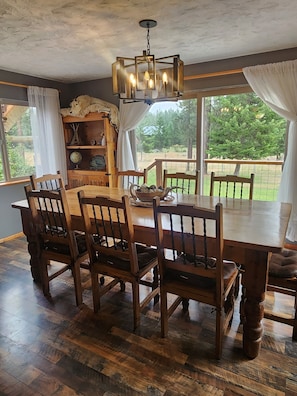 Un espace repas comprenant une grande table en bois, six chaises, un lustre et une vue sur un paysage verdoyant grâce à de grandes fenêtres.