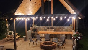 Une terrasse extérieure couverte, avec une table et des chaises en bois, un foyer et des guirlandes lumineuses.