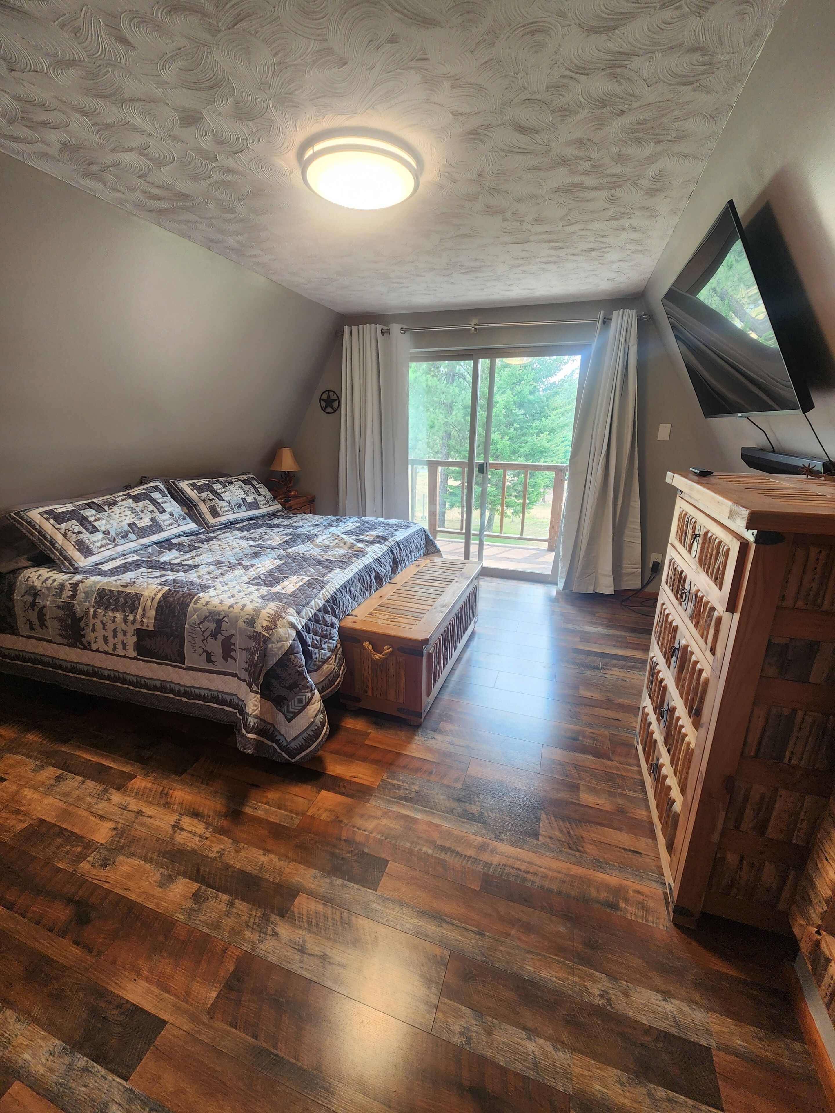 A bedroom with a large bed, a wooden chest of drawers, a television, and a balcony with curtains.
