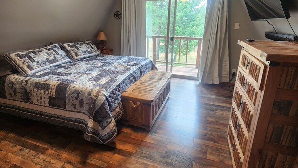 A bedroom with a large bed, a wooden chest of drawers, a television, and a balcony with curtains.
