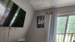 Une pièce comprenant une commode en bois, un téléviseur à écran plat fixé au mur et un tableau représentant des arbres.