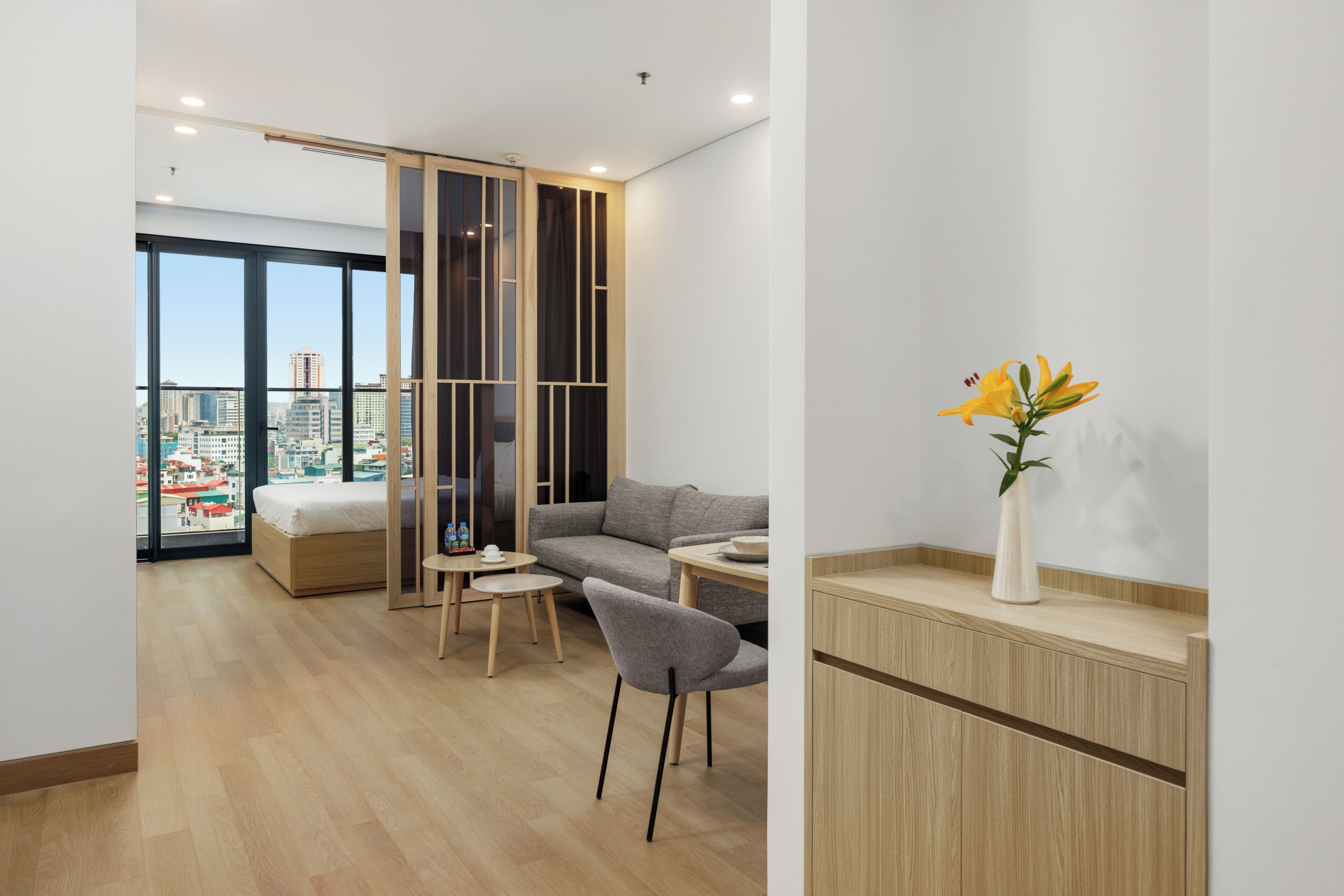 A modern hotel room with a bed, a gray sofa, a small dining table, a chair, and a vase with flowers.