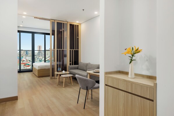 Una habitación de hotel moderna con una cama, un sofá gris, una pequeña mesa de comedor, una silla y un florero con flores.