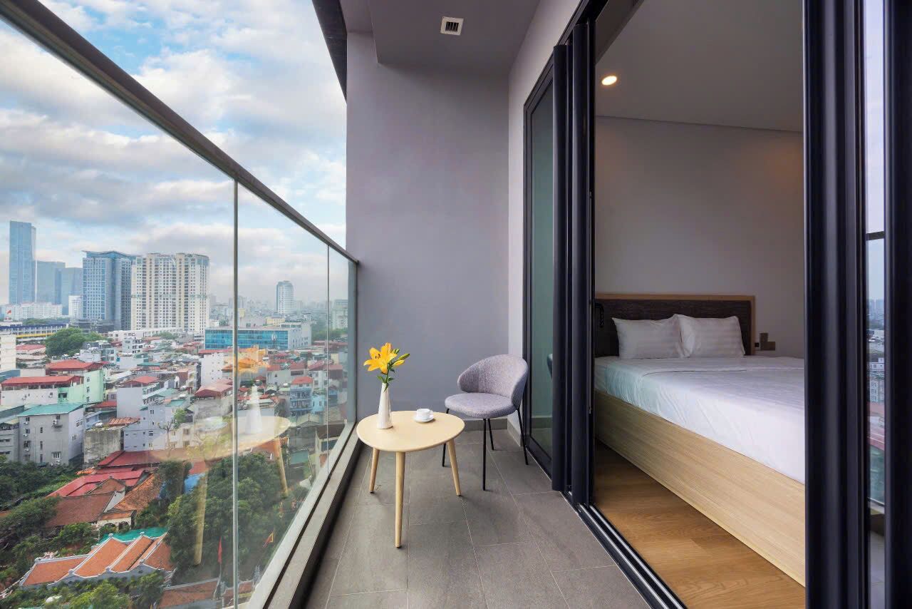 A modern bedroom with a large window, a bed, a small table with a vase, and a chair.