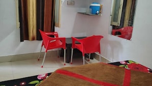 A room with a bed, two red chairs, a television, a mirror, and a small shelf with a blue container. - Nandishwar cottage (Mount Abu)