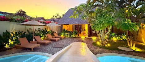 A pool area with lounge chairs and umbrellas, a thatched-roof structure, and tropical vegetation.