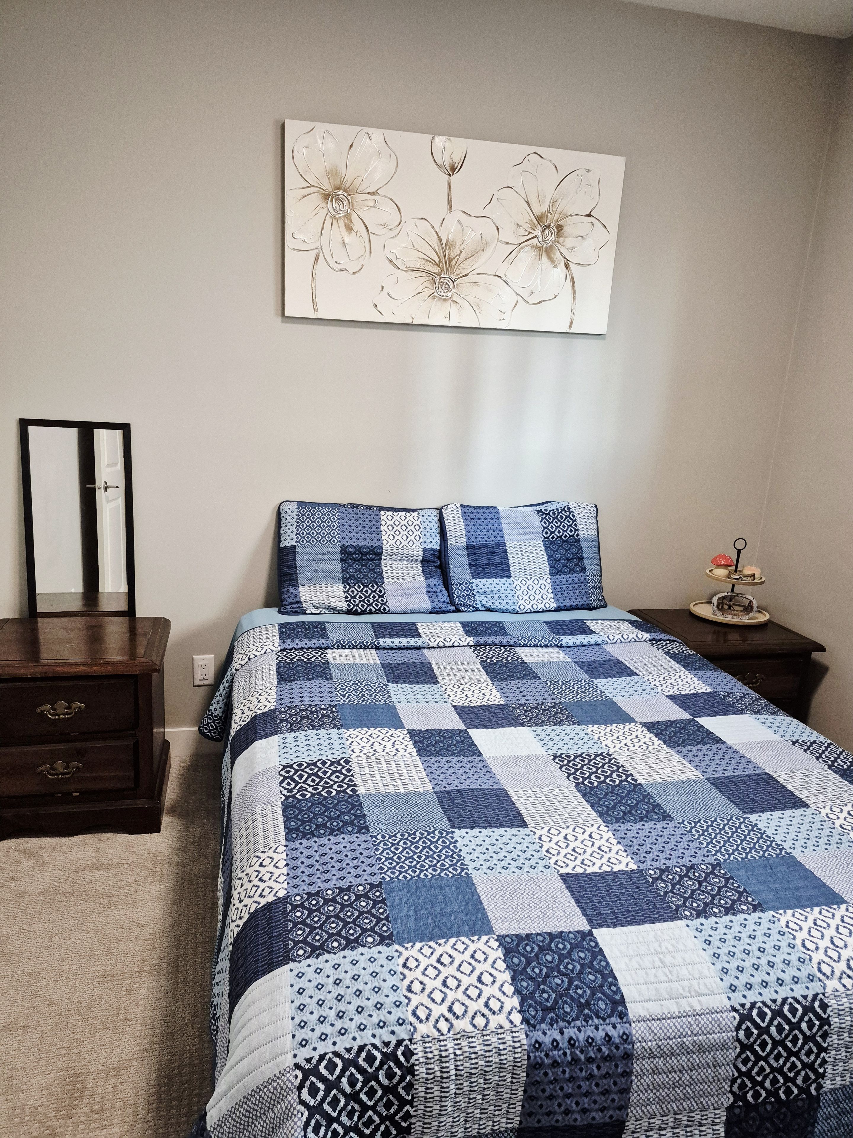 A bedroom with a bed, a dresser, a nightstand, a wall-mounted mirror, and a floral artwork.
