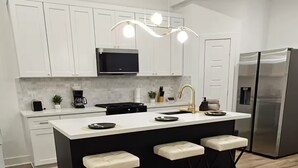 A modern kitchen with white cabinets, a central island with bar stools, a stainless steel refrigerator, and a hanging pendant light.