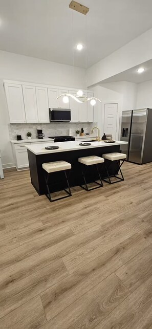 A modern kitchen with white cabinets, a central island with bar stools, a stainless steel refrigerator, and a hanging pendant light.