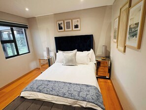 Een slaapkamer met een groot bed, twee nachtkastjes, een raam en kunstwerken in een lijst aan de muur.
