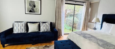 A bedroom with a bed, a dark blue sofa, a framed artwork, and a view through a window.