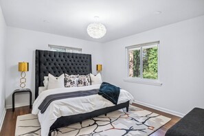 A bedroom with a tufted headboard, a large window, a bedside lamp, and a patterned rug. - Corporate Comfort | Stylish 3BR Extended Stay (Decatur)