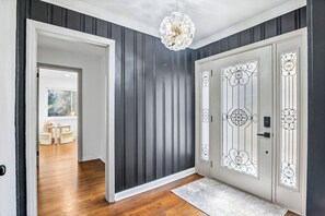 A hallway with dark vertical paneling, a white door with decorative glass panels, and a chandelier. - Corporate Comfort | Stylish 3BR Extended Stay (Decatur)