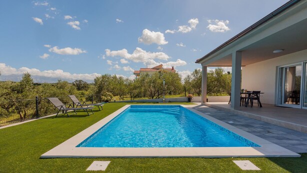 A swimming pool with a clear blue sky and clouds, surrounded by a well-maintained lawn and a building with a covered patio area. - Vacation home  by Interhome (Krneza)