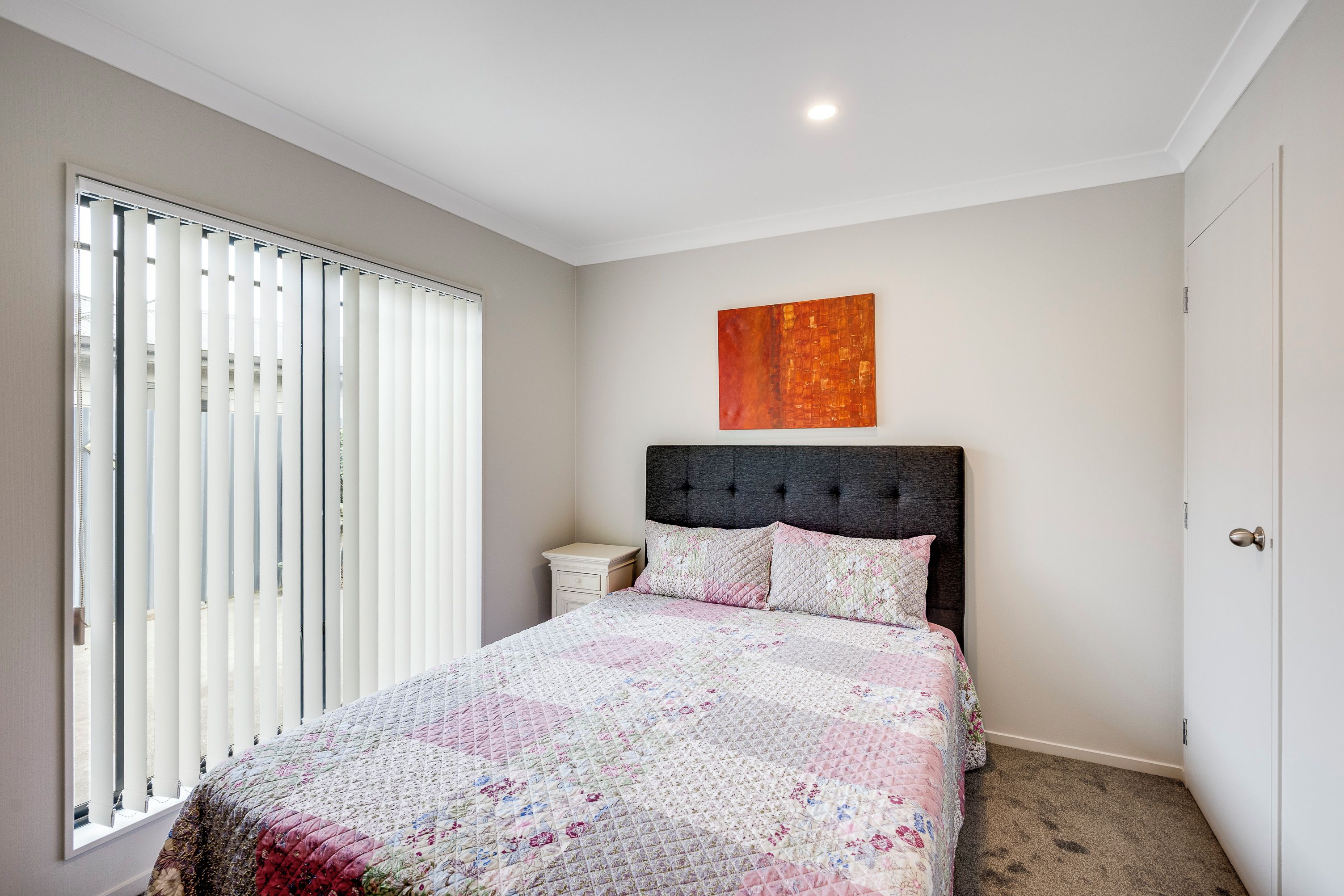 A bedroom with a bed, a nightstand, a window with blinds, and a painting on the wall.