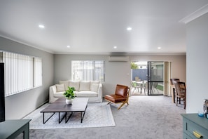 A modern living room with a sofa, a coffee table, and a brown leather chair. There is a sliding glass door leading to an outdoor patio area with a table and chairs.