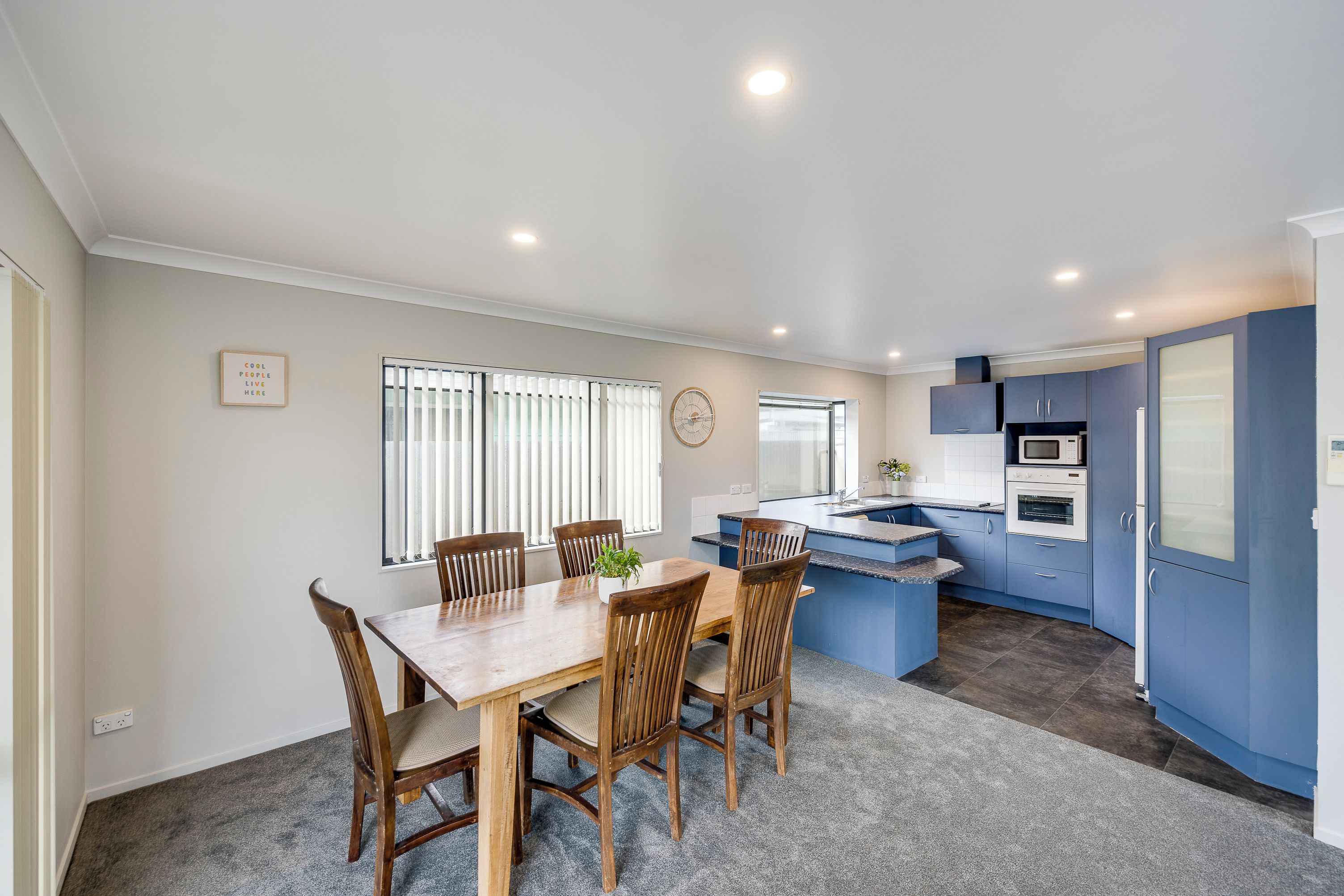A modern kitchen with blue cabinets, a wooden dining table, and a clock on the wall.