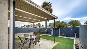 Un patio cubierto con una mesa y sillas, un jardín pequeño con césped artificial y una cerca azul.