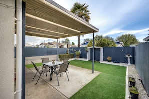 A covered patio area with a table and chairs, a small garden with artificial grass, and a blue fence.
