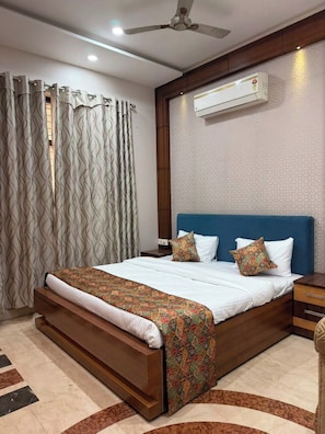 A bedroom with a bed, curtains, a ceiling fan, an air conditioning unit, and a patterned rug. - Rameshawram Sukoon Mansion (Varanasi)
