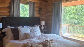 Et soveværelse i en træhytte med seng, puder, tæppe, et natbord, en lampe og udsigt til naturen.